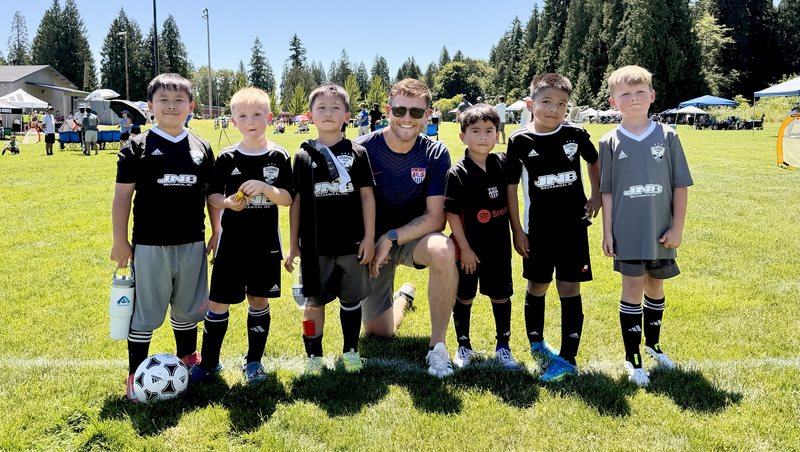 Coach Nick kneeling on a soccer field with six young players from a team he coached this summer